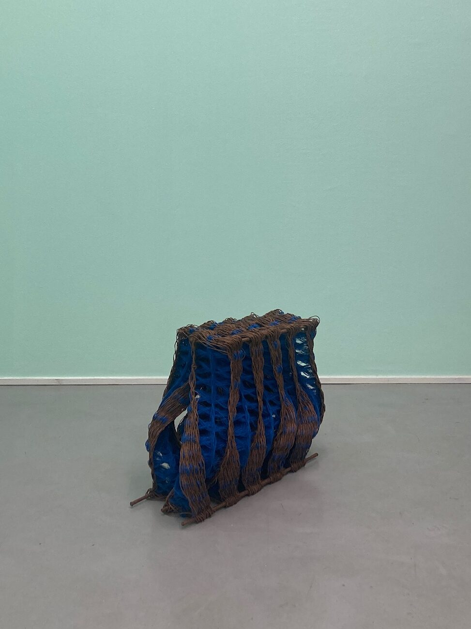 Blue and brown woven textile sculpture, placed on a gray concrete floor.