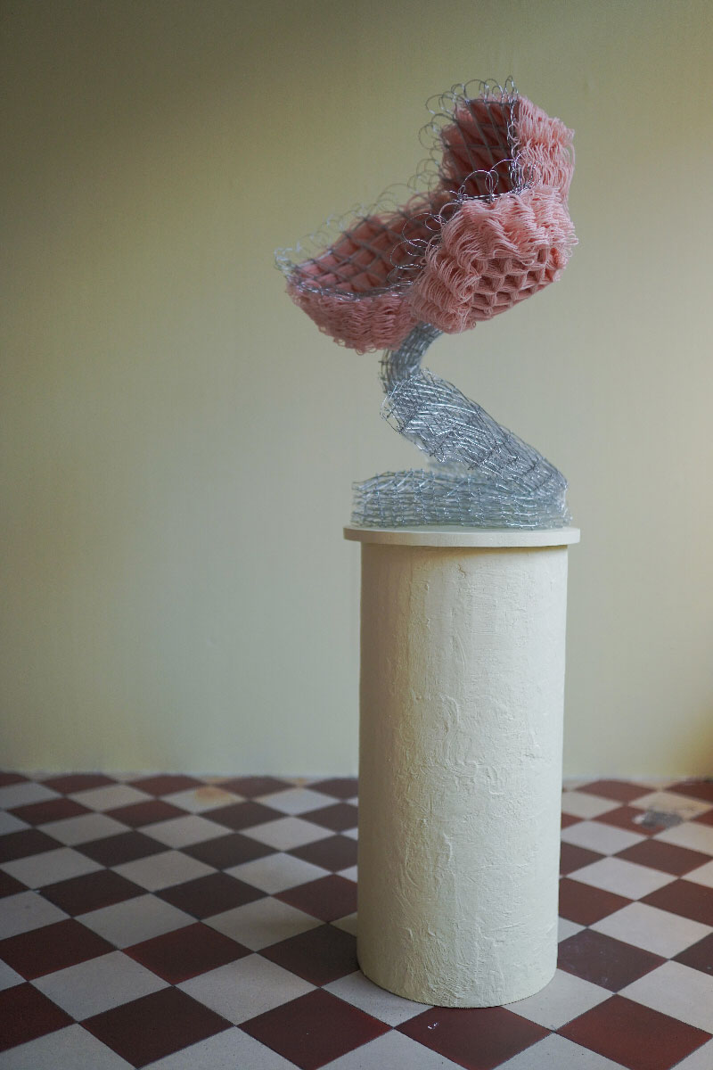 blomman. Abstract sculpture out of pink wool thread and steel wire on a yellow podium standing on a checked floor.