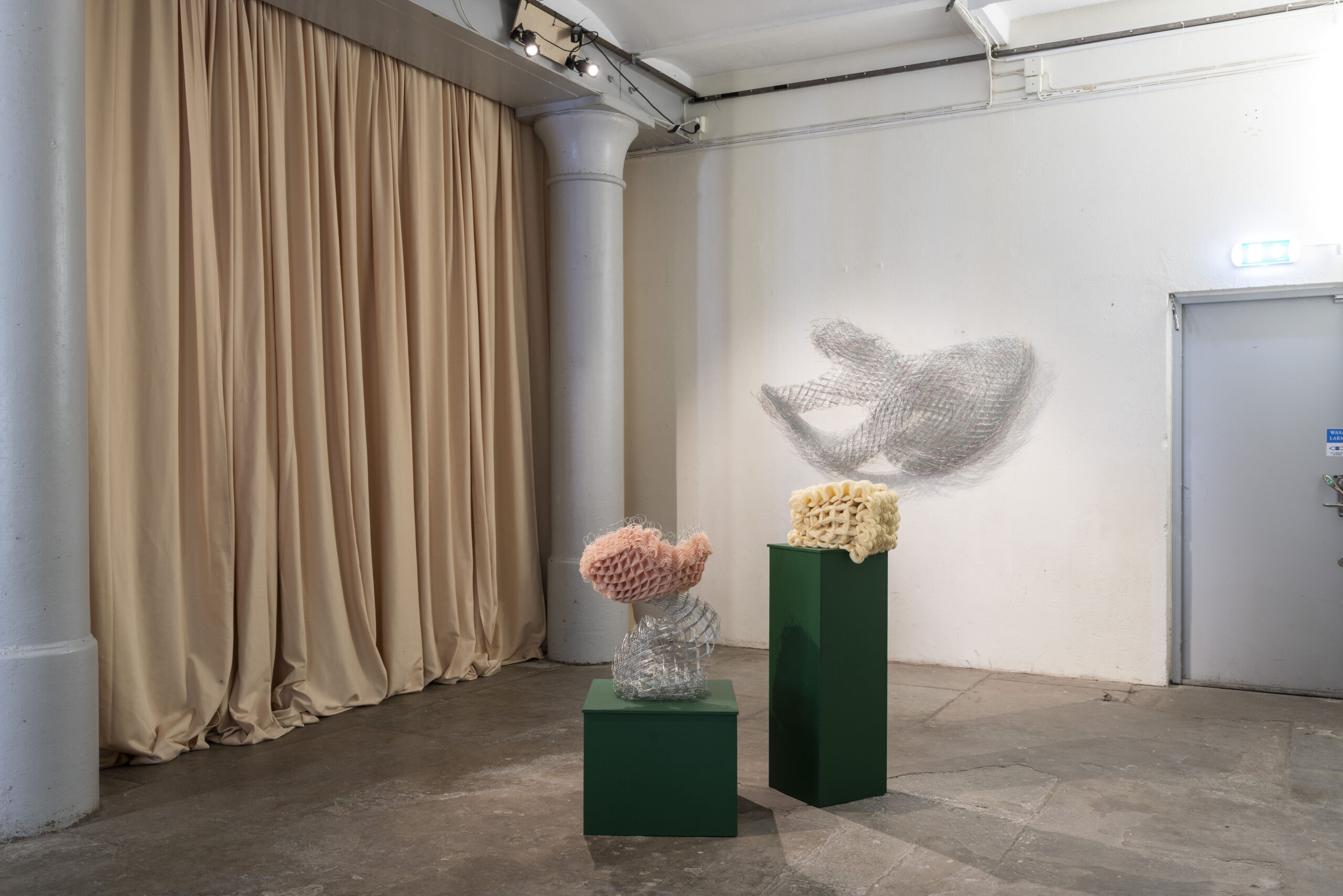 exhibition view. two sculptures in metal and textile placed on two dark green podiums in front of a metal sculpture hanging on a white wall.