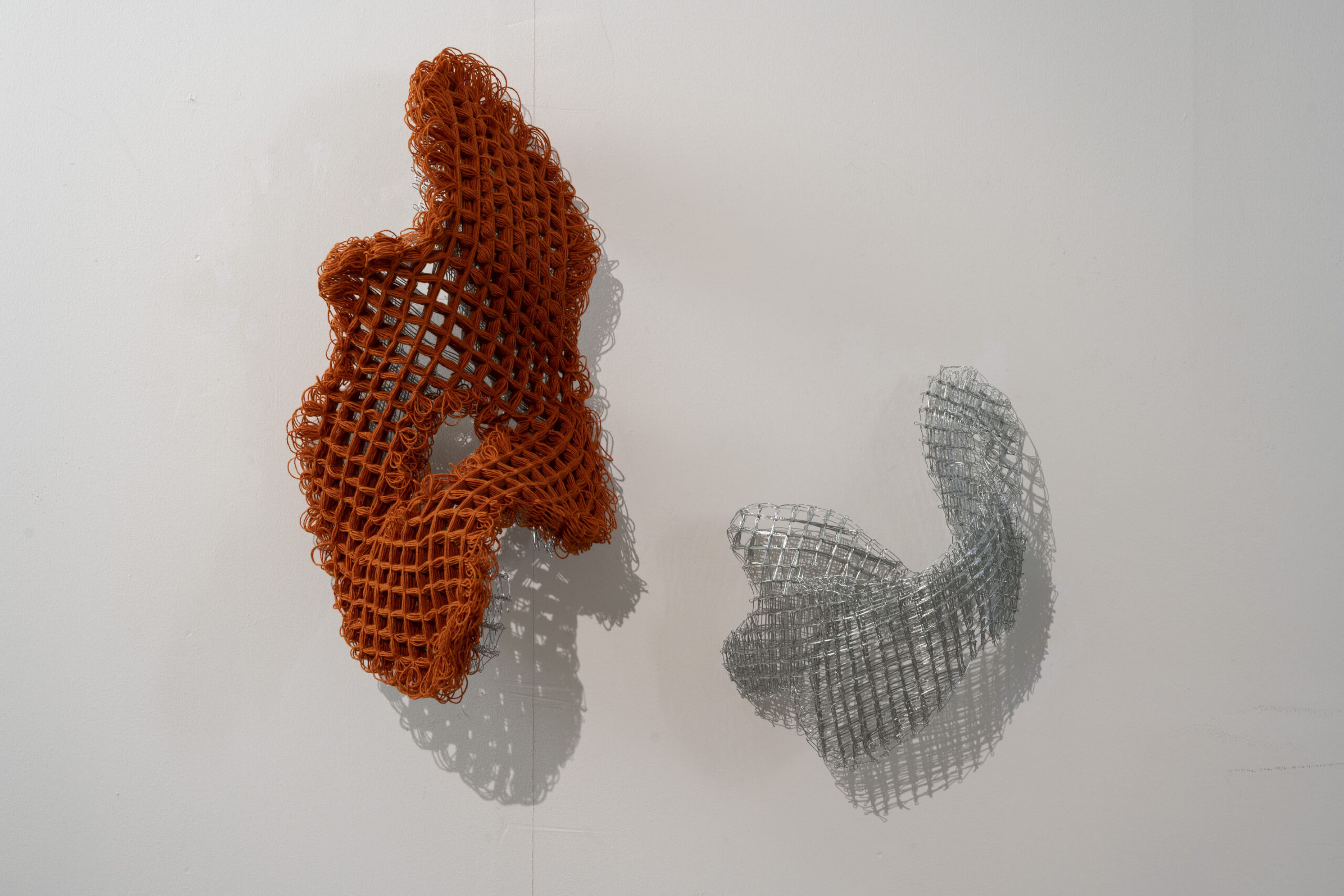 Two woven sculptures in orange wool yarn and metal on a white wall.