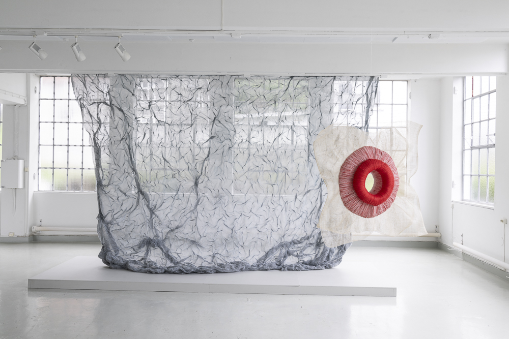 Red embroidery sculpture by Emilia Elfvik hanging in front of gray textile artwork by Kari Hjertholm.