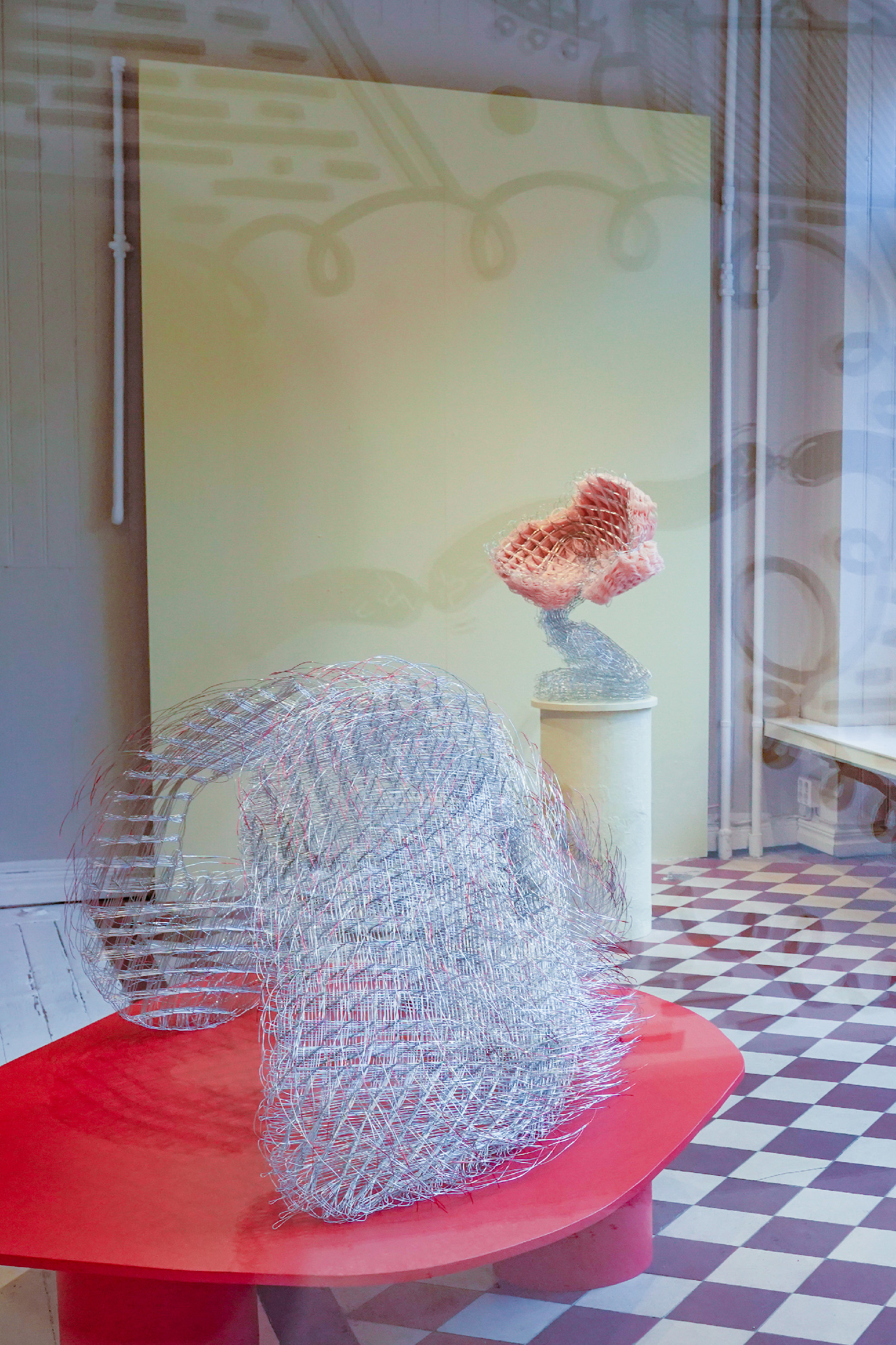 Exhibition view of Kontakt sökes. Metal sculpture on a red podium in the foreground, and a pink and steel coloured sculpture on a yellow podium in the background against a yellow wall. 